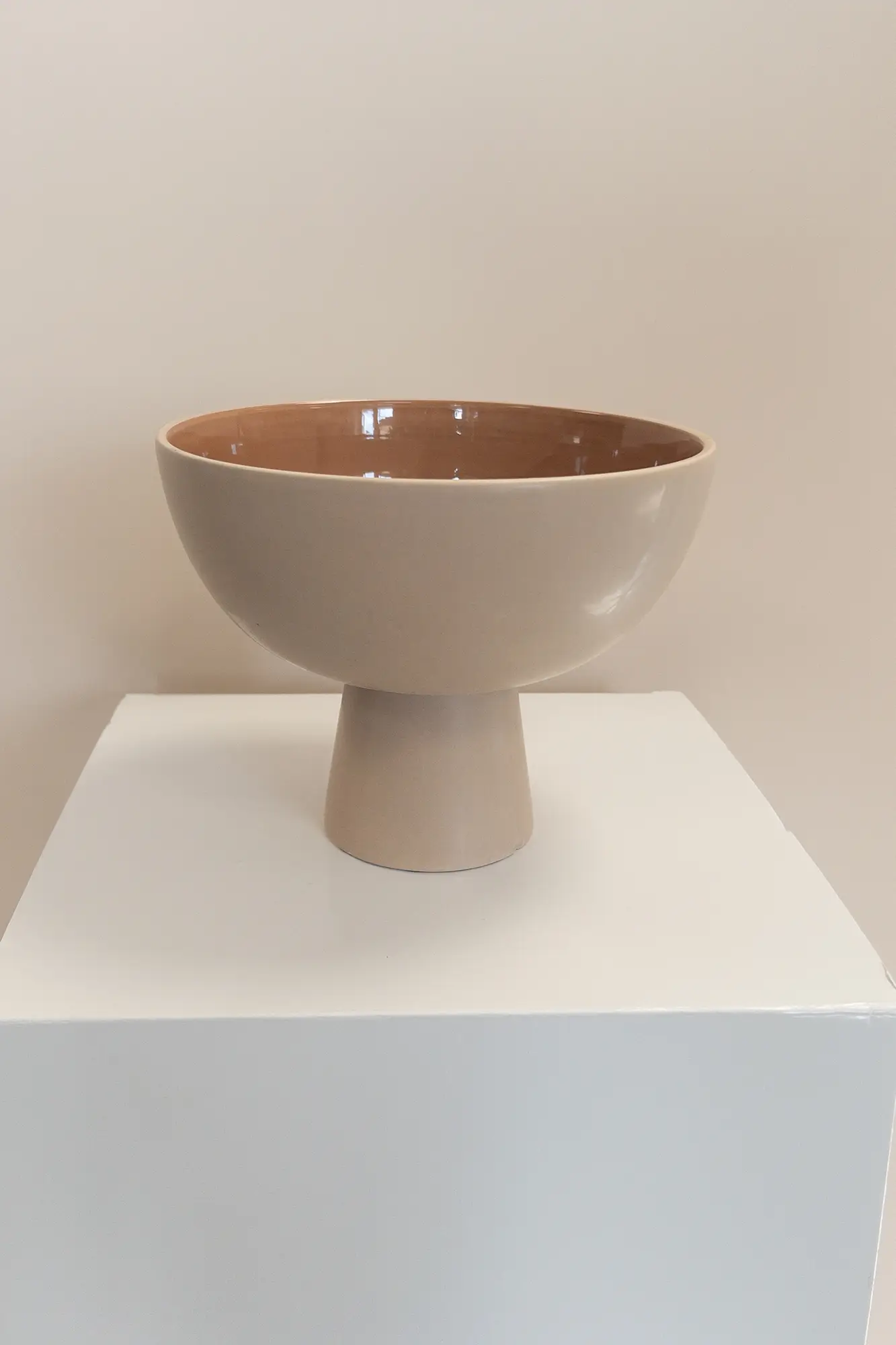 Eine beigefarbene Keramik-Sockelschale mit glänzender Innenseite von Dominique Kubicek steht auf einem weißen quadratischen Sockel, perfekt für moderne Hochzeitsfloristik, vor einem neutralen Hintergrund.