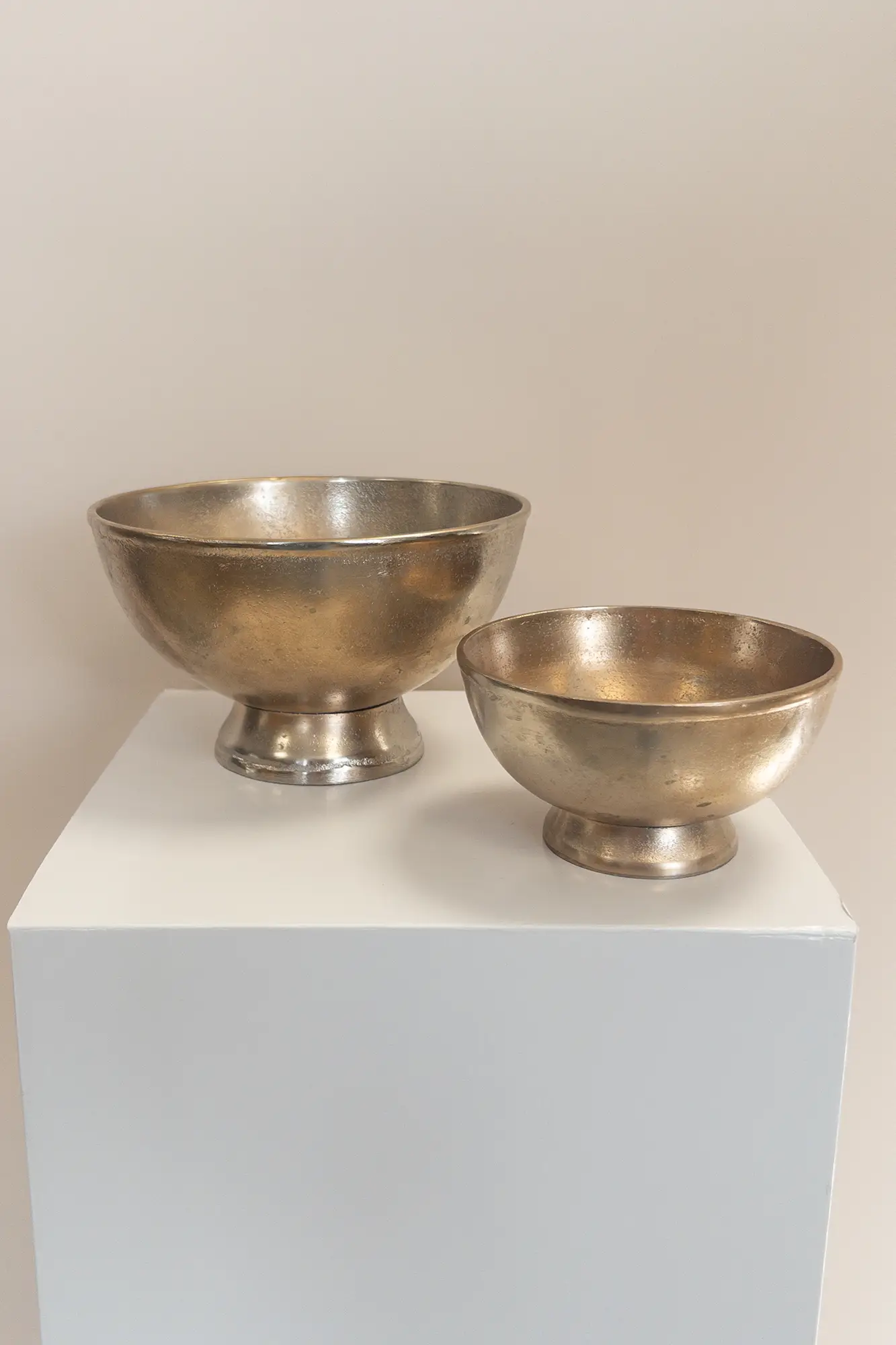 Zwei unterschiedlich große Metallic-Goldschalen stehen nebeneinander auf einem weißen Sockel vor einem schlichten beigen Hintergrund, perfekt für moderne Hochzeitsfloristik oder elegante Event & Floral Design Hochzeitsdekoration.