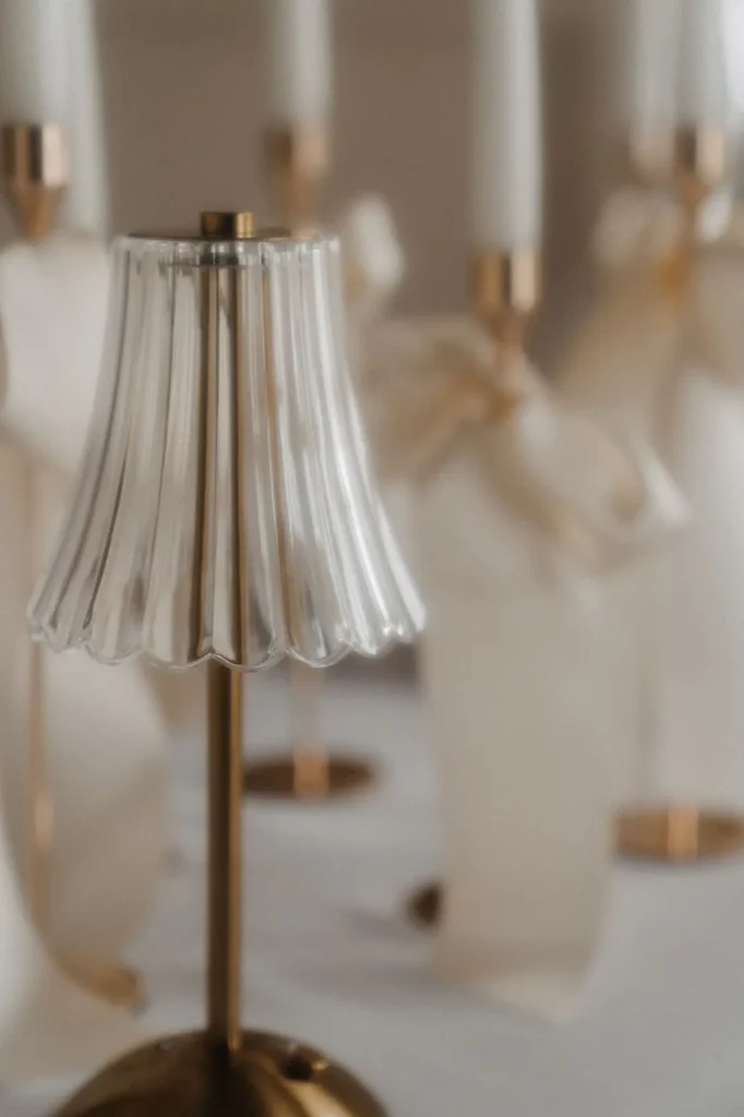 Eine kleine, elegante goldene Lampe mit einem klaren, gefalteten Glasschirm steht auf einem Tisch. Im unscharfen Hintergrund stehen hohe Kerzen mit goldenen Haltern und weißen Bändern, die den raffinierten Touch der modernen Hochzeitsfloristik von Dominique Kubicek widerspiegeln.
