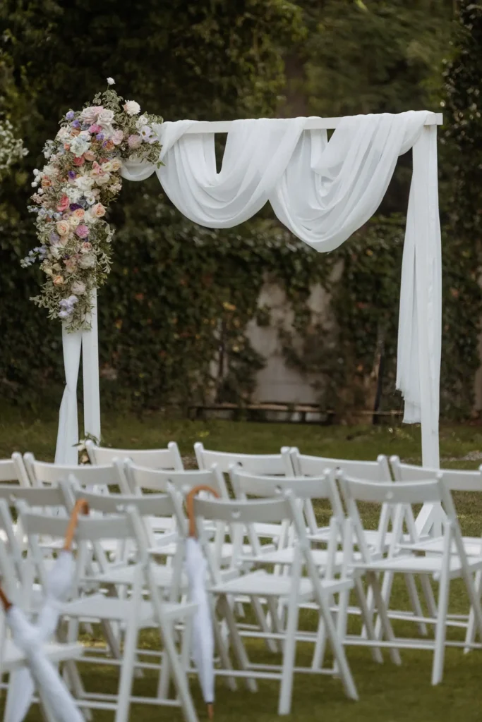 Ein weißer, mit durchsichtigem Stoff drapierter und mit pastellfarbenen Blumen geschmückter Hochzeitsbogen von Event & Floral Design Hochzeit steht vor Reihen von weißen Klappstühlen, die im Freien auf grünem Gras aufgestellt sind.