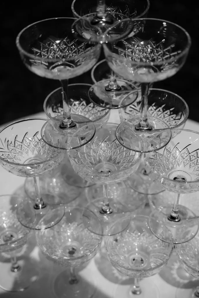 Ein Schwarz-Weiß-Foto einer Pyramide aus leeren, verzierten Kristallgläsern, die auf einem runden Tisch angeordnet sind, von oben gesehen. Perfekt für eine niederbayern Feier oder als Inspiration für moderne Hochzeitsfloristik mit seinem eleganten, symmetrischen Muster.