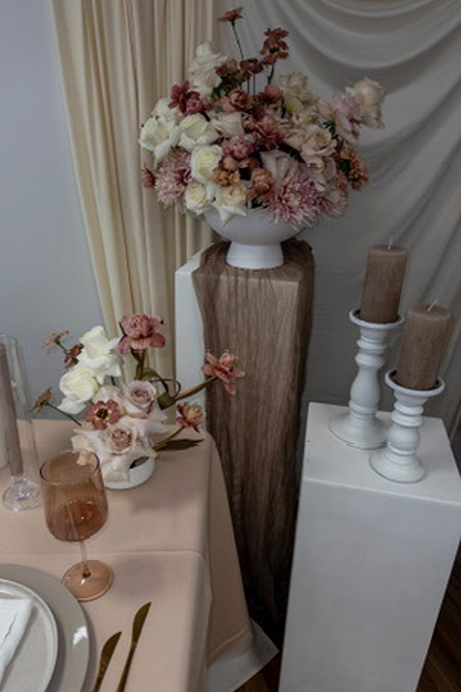 Ein Tisch mit rosafarbener und weißer Dekoration von Dominique Kubicek, mit einem großen Blumenarrangement auf einem Sockel, Kerzen in weißen Haltern und einem Weinglas - alles in sanften, neutralen Tönen auf drapiertem Stoff.