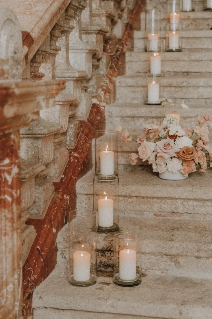 Eine rustikale Steintreppe, dekoriert mit brennenden weißen Kerzen in Glashaltern und einem Strauß aus rosa und cremefarbenen Blumen von Hochzeitsfloristik-Expertin Dominique Kubicek, schafft eine romantische und elegante Atmosphäre.