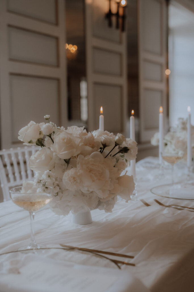 Ein sanft beleuchteter, eleganter Tisch mit weißen Blumen in einer Vase von Mondhaende Hochzeitsfloristik, vier brennenden weißen Spitzkerzen, goldenem Besteck und einem Glas Champagner - alles auf einem weißen Tischtuch in Niederbayern.