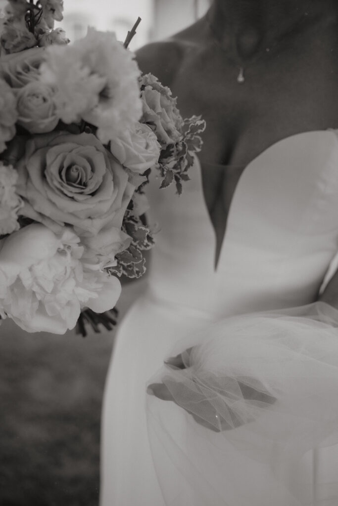 Ein Schwarz-Weiß-Foto einer Braut in einem trägerlosen Hochzeitskleid, die einen Strauß von mondhaende Event & Floral Design Hochzeit hält, mit einer zarten Halskette und einer Hand, die Tüll von ihrem Kleid sammelt - aufgenommen in Niederbayern.