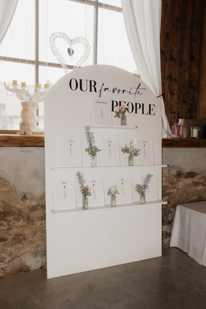 Ein weißer Sitzplan von Dominique Kubicek mit Hochzeitsfloristik mit Umschlägen und kleinen Blumenarrangements vor einer rustikalen Steinwand in Niederbayern, eingerahmt von weißen Vorhängen und in natürliches Licht getaucht.
