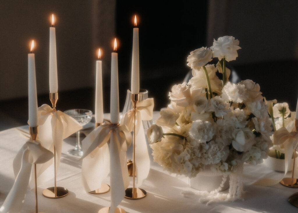 Ein Tisch, dekoriert mit fünf brennenden weißen Spitzkerzen in goldenen Haltern und Schleifen, daneben ein großer Strauß moderner Hochzeitsfloristik von mondhaende, schafft eine elegante, romantische Atmosphäre, perfekt für Hochzeiten in Niederbayern.