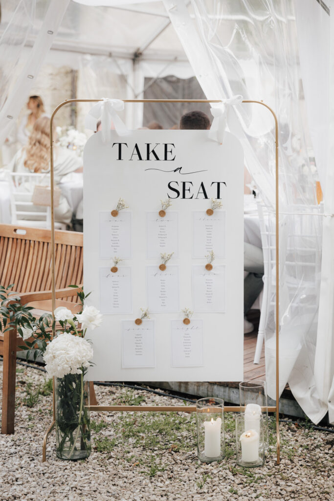 Ein Schild für den Sitzplan einer Hochzeit mit der Aufschrift "Take a Seat" (Nimm Platz), auf dem die Gästelisten mit Blumen angeheftet sind. Das Schild mit moderner Hochzeitsfloristik steht auf einem goldenen Rahmen neben weißen Blüten und Kerzen in einem hellen Zelt im Freien.