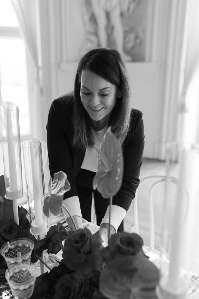 Eine Frau mit schulterlangen Haaren und Blazer arrangiert lächelnd Blumen und Kerzen auf einem dekorierten Tisch in einem hellen, eleganten Raum, der die moderne hochzeitsfloristik und Event & Floral Design Hochzeit von mondhaende präsentiert.