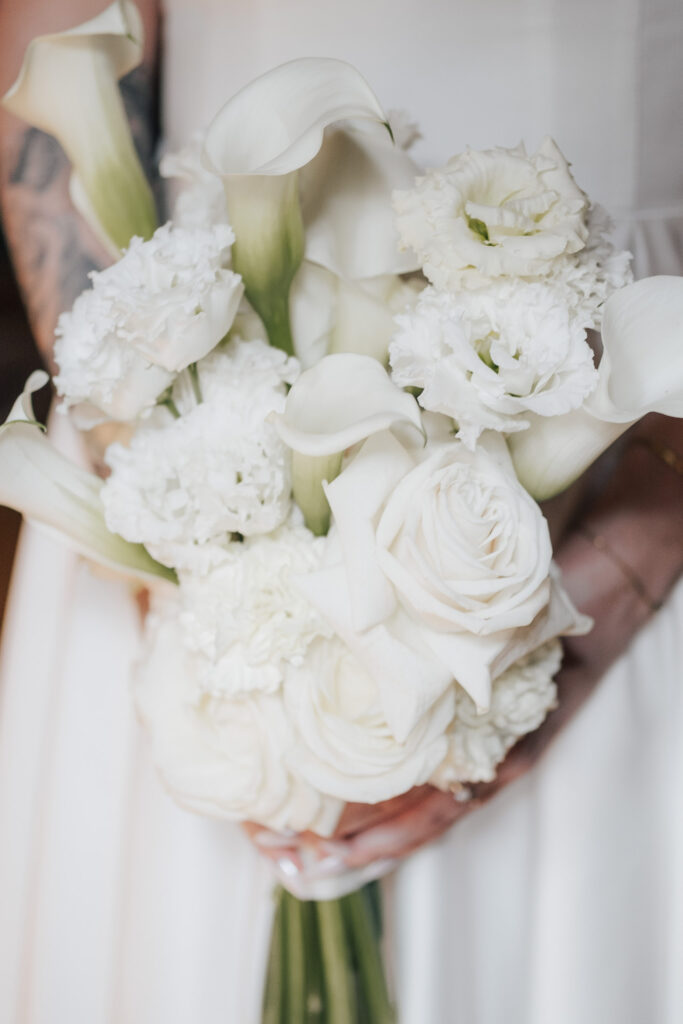 Eine Person in einem weißen Kleid hält einen Strauß weißer Blumen, darunter Rosen, Calla-Lilien und Nelken, die ein zartes und elegantes Arrangement bilden, das von Dominique Kubiceks Blumenkunst inspiriert ist.
