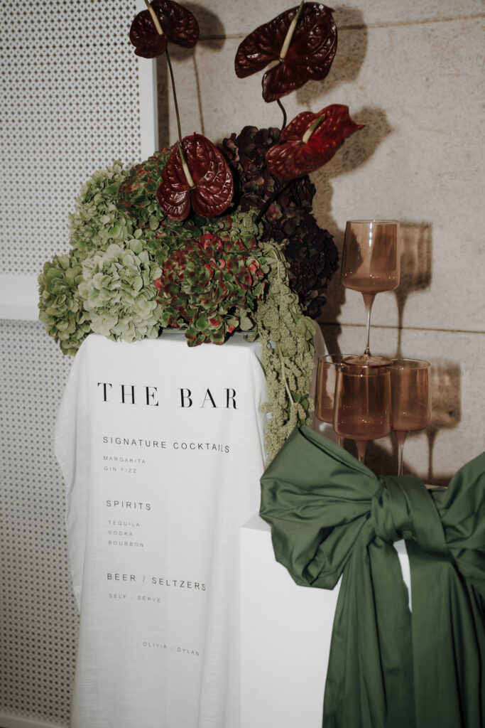 Eine Bar mit einer weißen Stoffkarte, auf der Cocktails, Spirituosen und Selters aufgelistet sind, ist über dem Tresen drapiert. Moderne Hochzeitsfloristik von Dominique Kubicek sorgt für rotes und grünes Flair, rosa Gläser und eine grüne Stoffdekoration runden den Look ab.