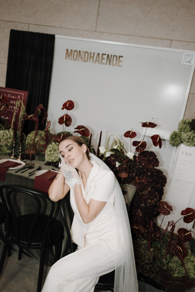 Eine Frau in weißem Outfit posiert mit geneigtem Kopf und der Hand vor dem Gesicht an einem elegant gedeckten Tisch, der mit roten und grünen Blumen geschmückt ist. Hinter ihr steht das Schild MONDHAENDE, das einen stilvollen Moment in diesem Dominique Kubicek niederbayern Lokal einfängt.