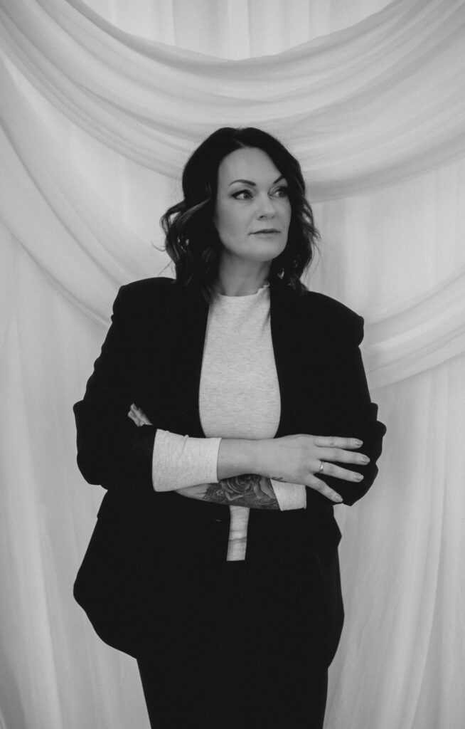 Eine Frau mit schulterlangem dunklem Haar steht mit verschränkten Armen da und trägt einen dunklen Blazer über einem hellen Hemd. Mit ernster Miene und einer sichtbaren Tätowierung am Unterarm steht sie vor einem drapierten weißen Stoff - perfekt für eine Event & Floral Design Hochzeit.