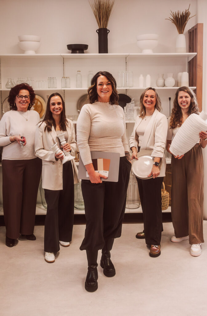 Fünf Frauen stehen in einem stilvollen Raum mit Vasen- und Glasregalen, lächeln und halten Teller und Vasen. Gekleidet in abgestimmte, neutrale Töne, verkörpern sie die Eleganz der modernen Hochzeitsfloristik in Niederbayern.
