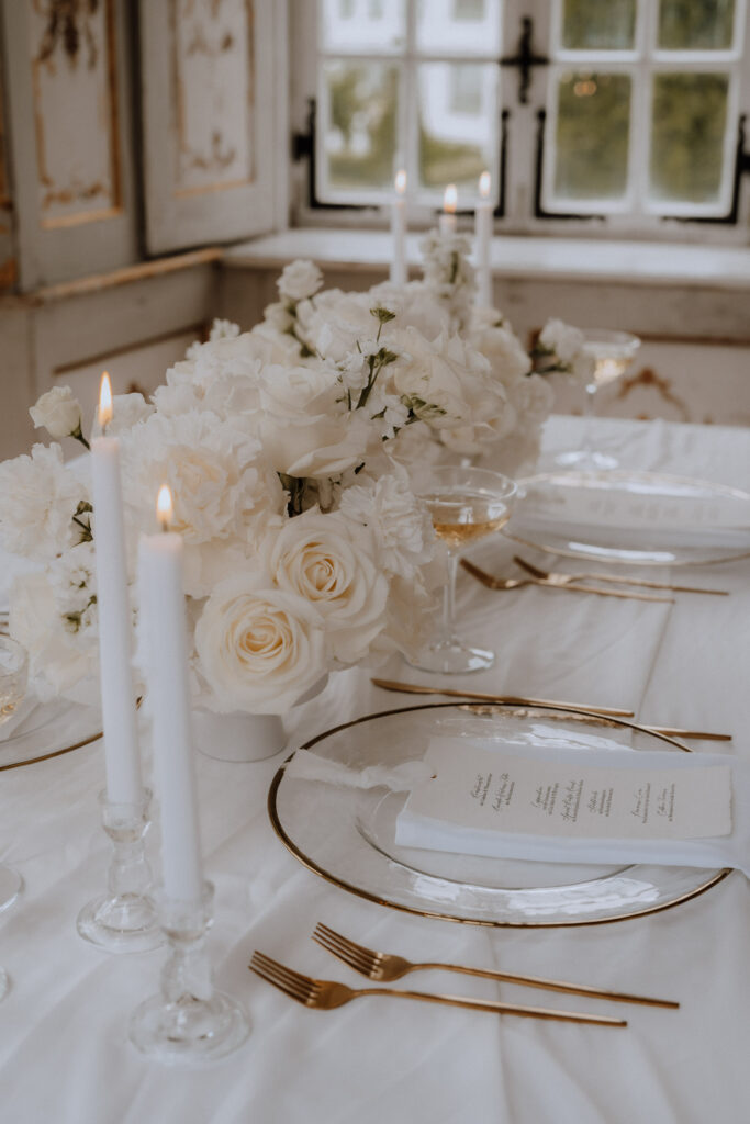 Elegant gedeckter Tisch mit goldenem Besteck, durchsichtigen Tellern mit Speisekarten, weißem Blumenschmuck von Dominique Kubicek moderne hochzeitsfloristik und brennenden weißen Kerzen auf einem weißen Tischtuch am Fenster.