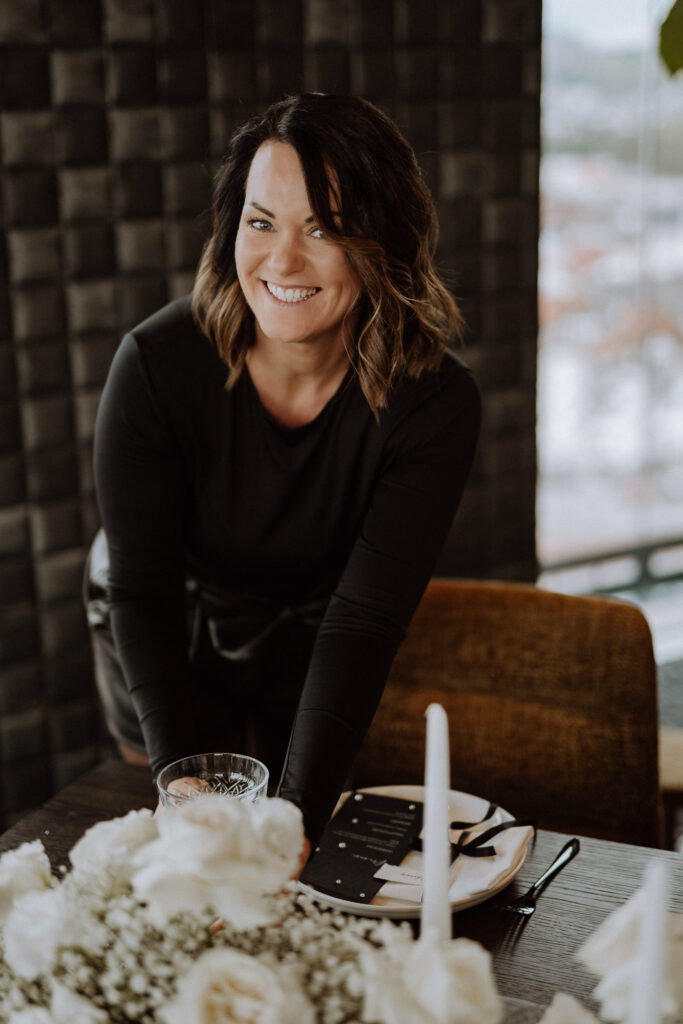 Eine lächelnde Frau mit schulterlangen braunen Haaren und schwarzem Oberteil beugt sich über einen mit weißen Blumen von Dominique Kubicek, Event & Floral Design Hochzeit, gedeckten Esstisch in einem modernen, sanft beleuchteten Raum.