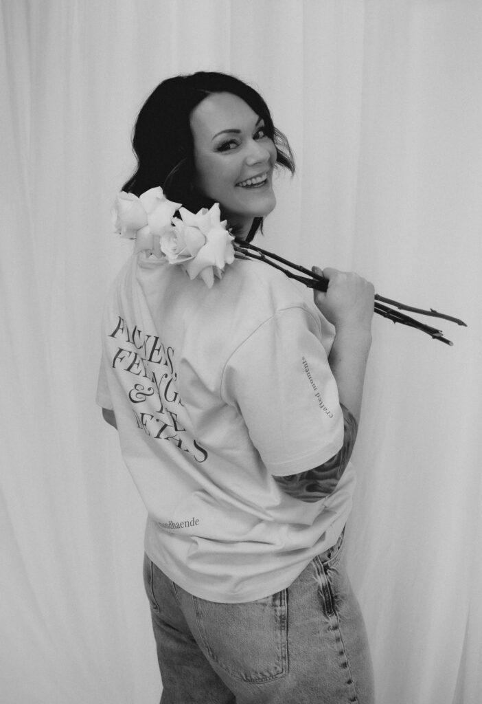 Eine Frau mit dunklen Haaren lächelt über ihre Schulter und hält drei Rosen, die auf moderne Hochzeitsfloristik hindeuten. Sie trägt ein helles T-Shirt mit Text auf dem Rücken und eine Jeans und steht auf diesem Schwarz-Weiß-Foto aus Niederbayern vor einem weißen Hintergrund.