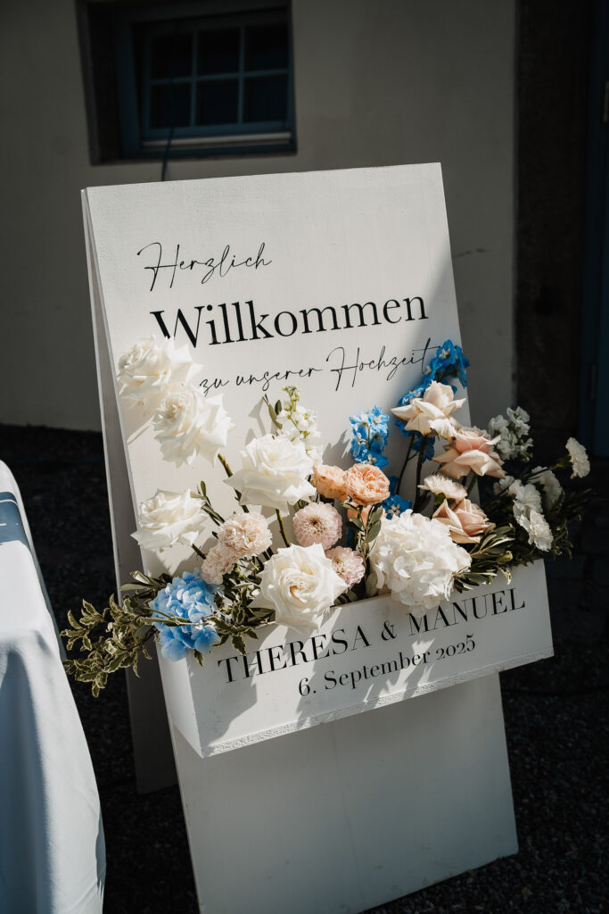 Ein weißes Hochzeits-Willkommensschild von Dominique Kubicek Event & Floral Design Hochzeit, mit moderner Hochzeitsfloristik in Weiß, Pfirsich und Blau, begrüßt die Gäste: Herzlich Willkommen zu unserer Hochzeit. Theresa & Manuel, 6. September 2023.