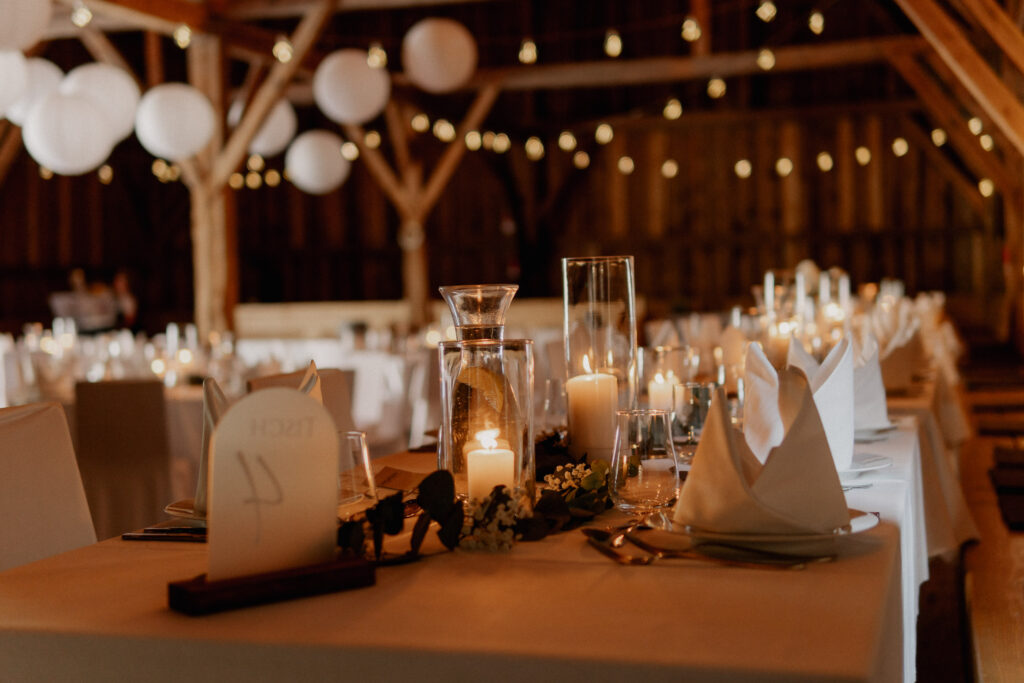 Elegant gedeckter Banketttisch für eine Veranstaltung in einer rustikalen Holzhalle, mit weißer Bettwäsche, Gläsern, Kerzen und Lichterketten. Hochzeitsfloristik von Dominique Kubicek verleiht dieser intimen niederbayerischen Feier Charme und Stil.