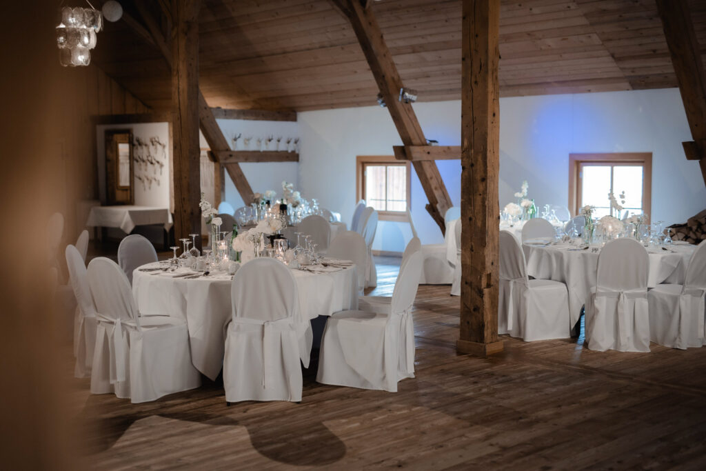 Ein rustikaler Veranstaltungssaal in Niederbayern mit Holzbalken, runden Tischen mit weißen Tüchern und modernen Hochzeitsfloristik-Mittelstücken schafft eine einladende, formelle Atmosphäre, die perfekt für jede Feier ist.