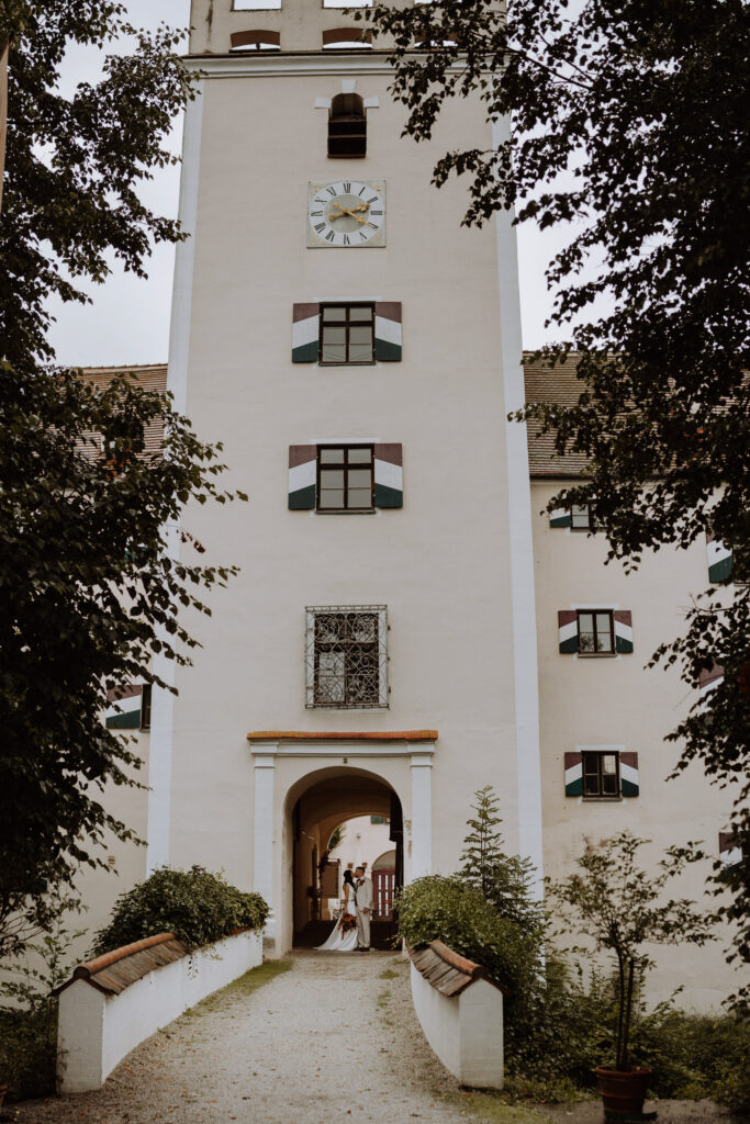 Eine Braut und ein Bräutigam stehen zusammen unter einem gewölbten Eingang eines hohen, beigen Gebäudes in Niederbayern mit grünen und roten Fensterläden, moderner Hochzeitsfloristik von Dominique Kubicek und einer großen Uhr darüber, umgeben von Bäumen und einem Kiesweg.