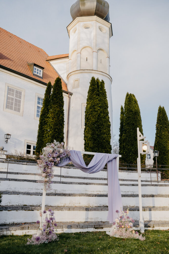 Ein mit lilafarbenem Stoff drapierter und mit lila und weißen Blumen geschmückter Hochzeitsbogen zeigt moderne Hochzeitsfloristik auf Steinstufen, mit hohen grünen Bäumen und einem weißen Gebäude mit rundem Turm im Hintergrund.