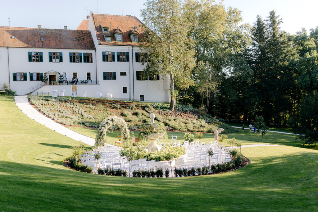Bei einer Trauung im Freien stehen weiße Stühle in einem Kreis mit moderner Hochzeitsfloristik von mondhaende vor einem großen weißen historischen Gebäude, umgeben von grünem Rasen und Bäumen unter einem klaren Himmel.