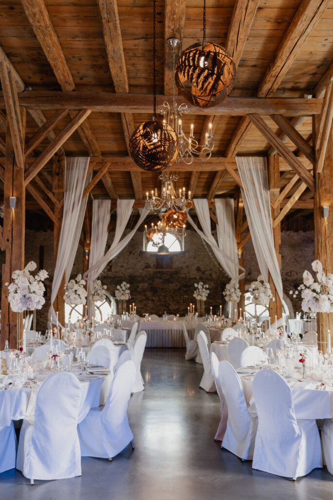 Ein rustikaler, eleganter Hochzeitssaal mit Holzbalken und weiß drapierten Stoffen von Event & Floral Design Hochzeit bietet runde Tische mit hochzeitsfloristischen Tafelaufsätzen, Kronleuchter und warme Beleuchtung für eine romantische Atmosphäre.