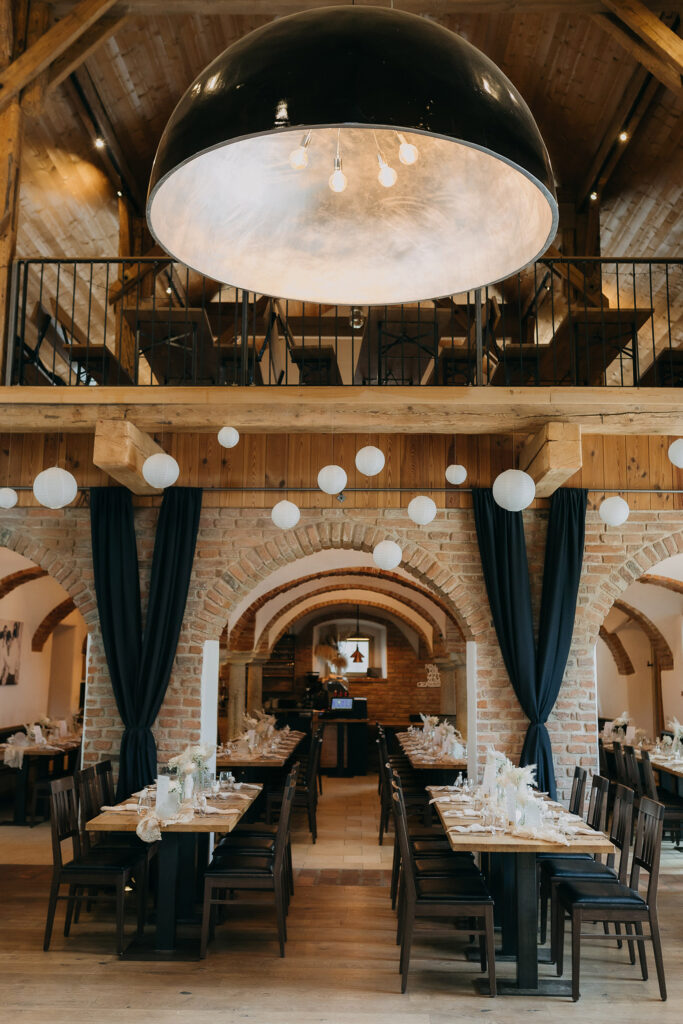 Ein rustikaler Speisesaal mit Holzdecken, freiliegenden Backsteinbögen, Tischreihen für das Essen und eleganten Akzenten von Event & Floral Design Hochzeit. Darüber hängen weiße Laternen und eine schwarze Lichtkuppel, während ein Balkon mit Geländer die Szene überblickt.