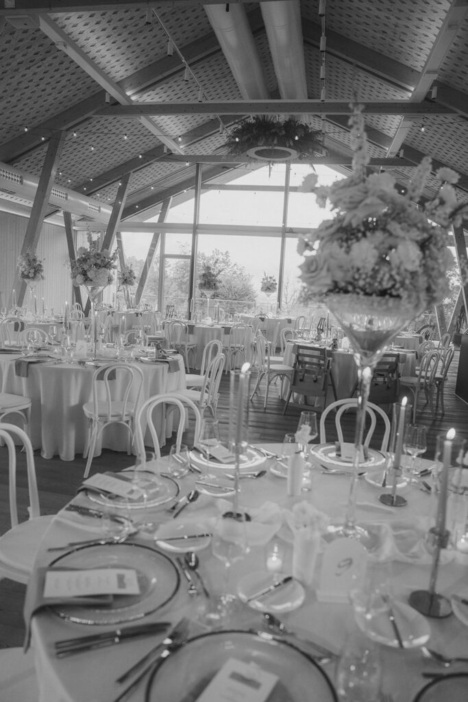 Ein Schwarz-Weiß-Foto eines eleganten Bankettsaals für eine Veranstaltung von Dominique Kubicek, mit moderner Hochzeitsfloristik mit hohen Blumenkränzen, runden Tischen, Kerzen und großen Fenstern unter einer gewölbten Decke.