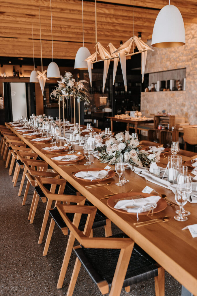 Ein langer, hölzerner Esstisch für eine elegante Veranstaltung von Dominique Kubicek, dekoriert mit Hochzeitsfloristik, goldenen Platzdeckchen und Origami-Kranichen. Moderne Pendelleuchten vervollständigen das stilvolle Event & Floral Design Hochzeit-Ambiente.