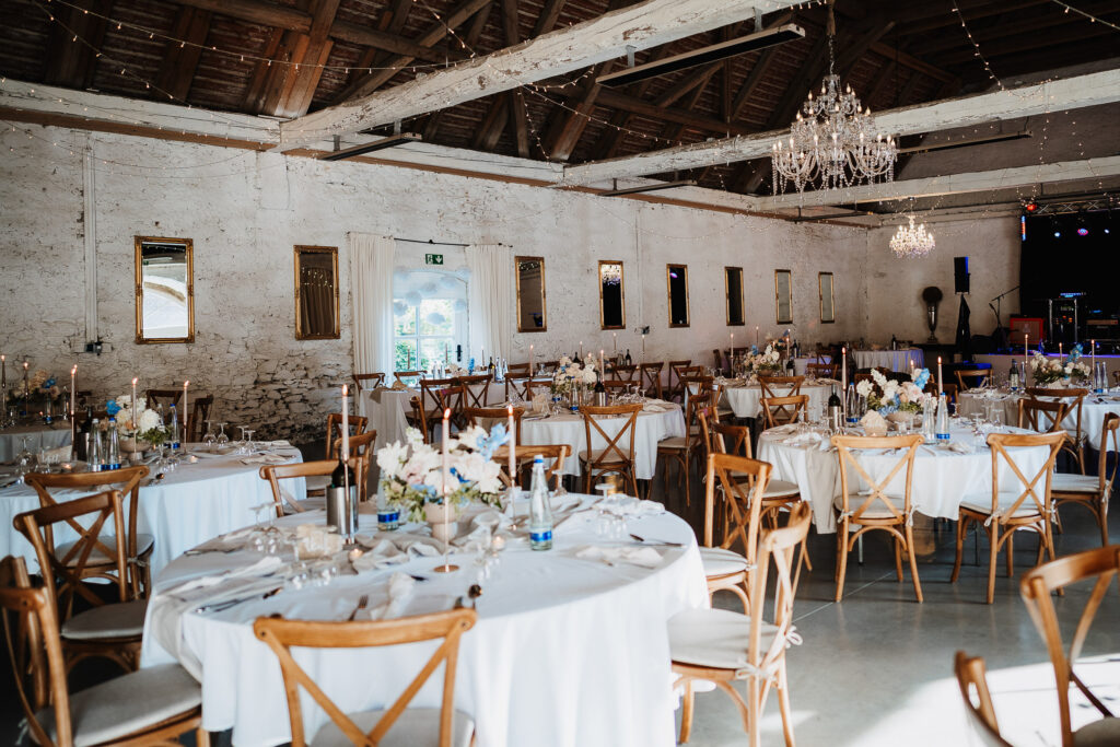 Ein rustikaler Hochzeitssaal mit Holzbalken, Kronleuchtern und Lichterketten. Die runden Tische sind elegant mit Event & Floral Design Hochzeits-Mittelstücken und Holzstühlen gedeckt. Der Raum hat eine gemütliche, einladende Atmosphäre.