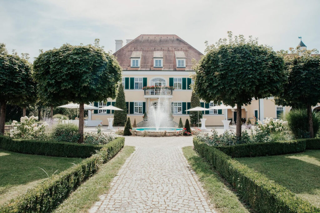 Ein kopfsteingepflasterter Weg führt zu einer gelben Villa mit grünen Fensterläden, einem Balkon und einem zentralen Brunnen - eine idyllische Kulisse, die durch Dominique Kubiceks hochzeitsfloristik und Event & Floral Design Hochzeit mit gestutzten Hecken und ordentlich geschnittenen Bäumen noch verstärkt wird.