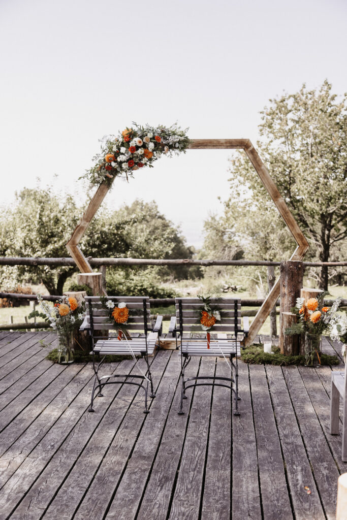 Zwei Metallstühle mit orangefarbenen Blumendekorationen von Dominique Kubicek sitzen auf einer Holzterrasse vor einem großen sechseckigen Holzbogen, der mit mondhaende hochzeitsfloristik geschmückt ist, umgeben von üppigem Grün und Bäumen in einem Außenbereich.