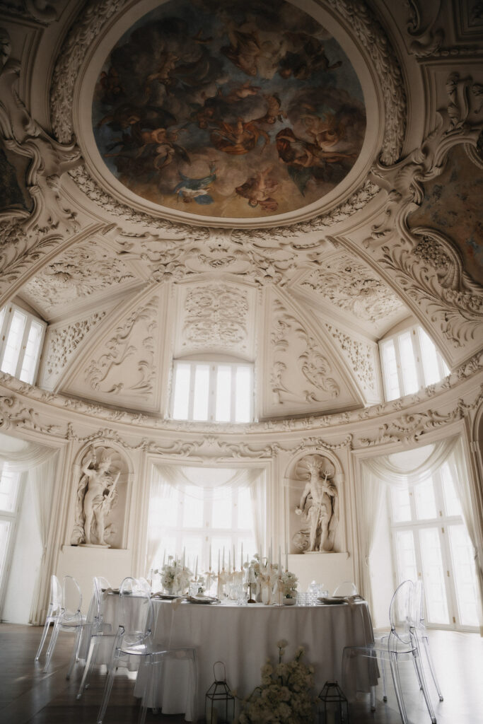 Ein prächtiger, verzierter Raum mit kunstvollen weißen Stuckleisten, großen Fenstern, einer bemalten Kuppeldecke und Statuen. Unter der Decke befindet sich ein runder Tisch mit eleganter moderner Hochzeitsfloristik aus Niederbayern und transparenten Stühlen.