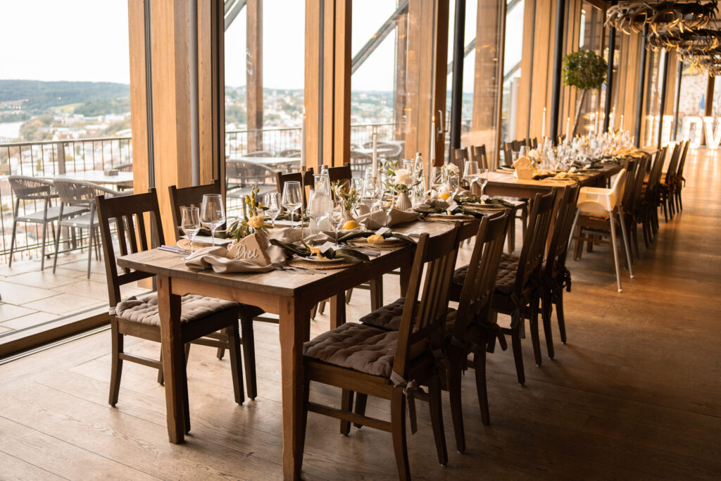 Elegante, mit Tellern, Gläsern und Servietten gedeckte Esstische stehen an großen Fenstern in einem sonnendurchfluteten, modernen Restaurant mit Holzböden und Blick nach draußen - wunderschön ergänzt durch moderne Hochzeitsfloristik von Dominique Kubicek.