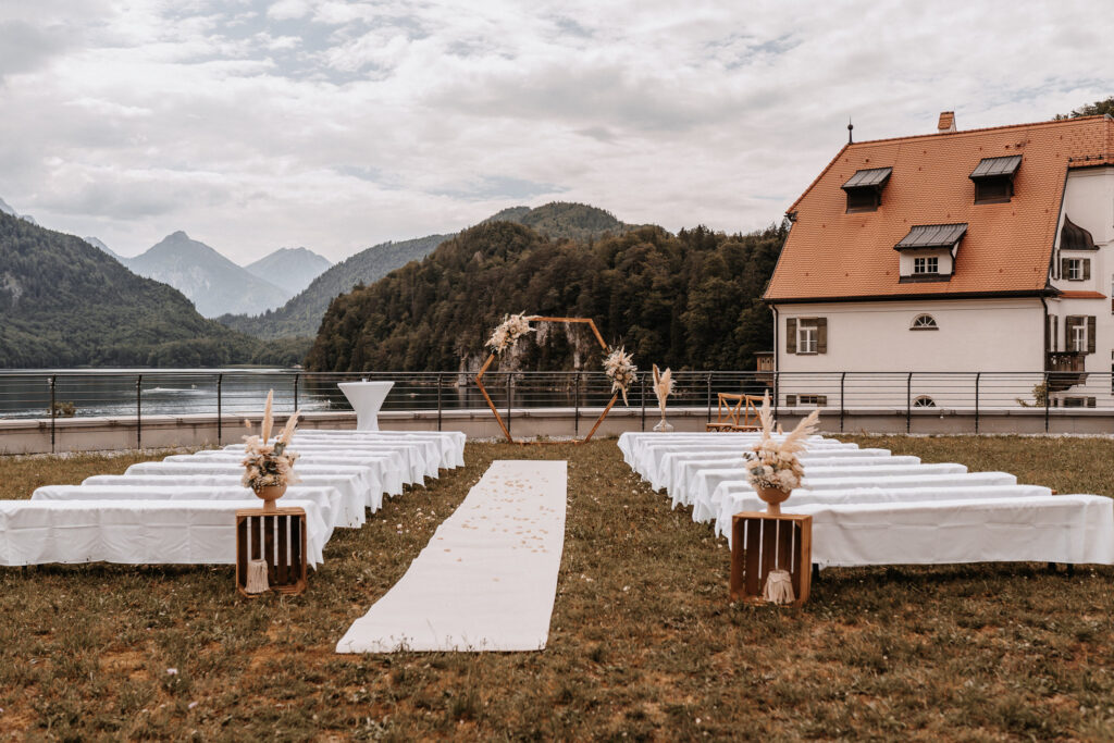 Hochzeitszeremonie im Freien von mondhaende Event & Floral Design Hochzeit, mit weiß gedeckten Bänken, einem blumengeschmückten Holzbogen und einem weißen Gangläufer auf einer Wiese mit Blick auf einen See, Berge und ein rotes Haus in Niederbayern.