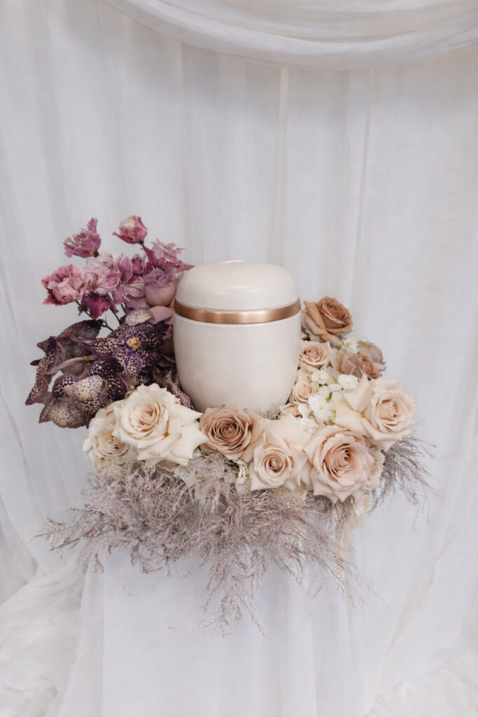 Eine weiße Urne mit einem goldenen Band ist umgeben von pastellfarbenen Rosen, violetten Blumen und Pampasgras, die alle auf einem weiß drapierten Stoffhintergrund arrangiert sind - eine elegante Note der Hochzeitsfloristik von Dominique Kubicek.