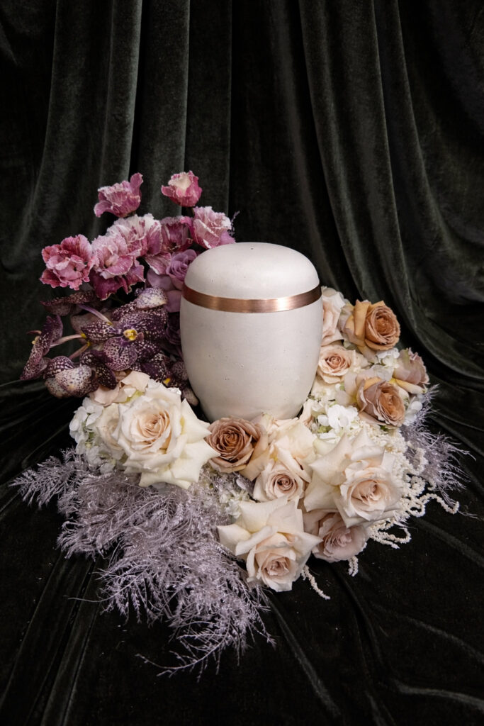 Eine weiße Kremationsurne mit Bronzeband ist umgeben von hellen Rosen, lavendelfarbenen Orchideen und silbernem Laub - alles arrangiert auf einem dunkelgrünen Samthintergrund. Eine exquisite Hochzeitsfloristik, inspiriert von mondhaende in Niederbayern.