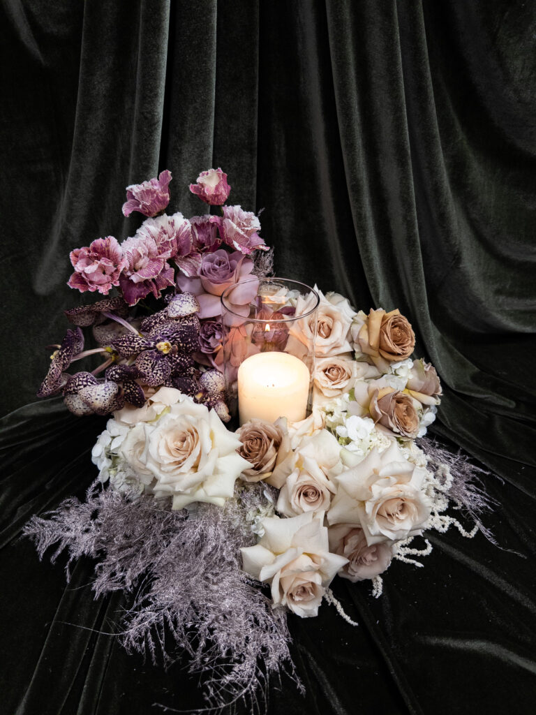 Eine brennende Kerze in einem Glashalter ist umgeben von cremefarbenen, beigen und violetten Rosen, Orchideen und gefiederten Blättern - ein elegantes Beispiel für moderne Hochzeitsfloristik von Dominique Kubicek auf dunklem, samtigem Stoff in Niederbayern.