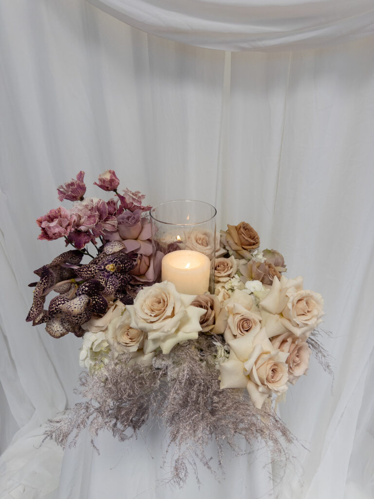 Ein Blumenarrangement von Dominique Kubicek Event & Floral Design Hochzeit mit hellen Rosen, violetten Orchideen und zartem Grün umgibt eine brennende Kerze in einem Glas, die auf weißem Stoff drapiert ist.