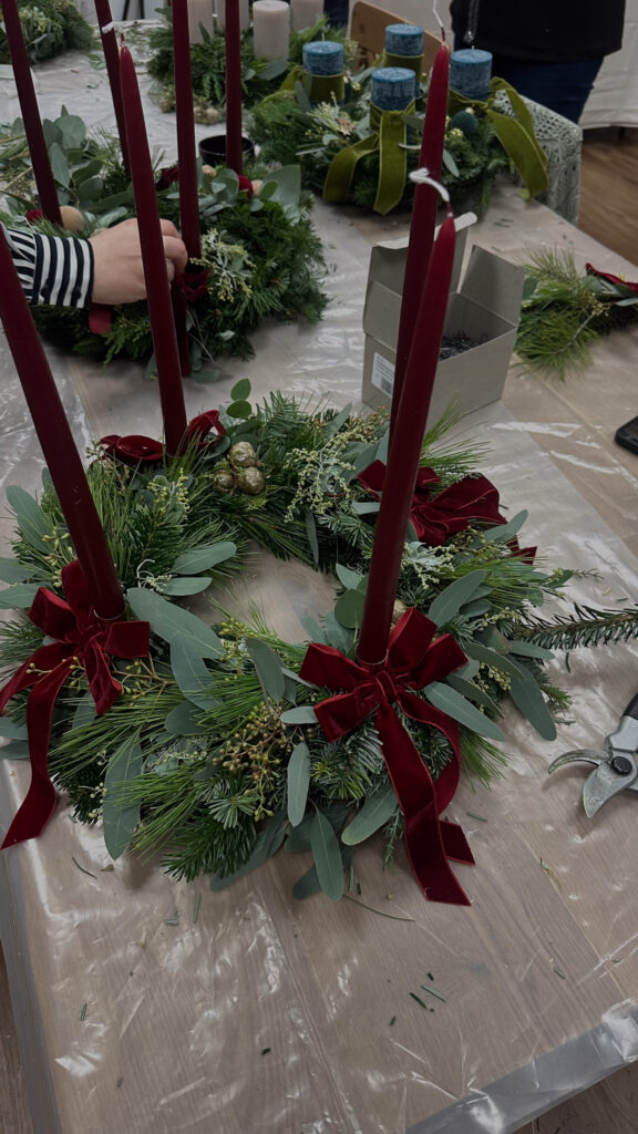 Eine Person arrangiert einen festlichen Kranz mit Grünzeug, roten Samtschleifen und vier hohen roten Kerzen auf einem mit Plastik abgedeckten Tisch, der kreative Hochzeitsfloristik zeigt. Andere Kränze und Materialien sind im Hintergrund zu sehen.