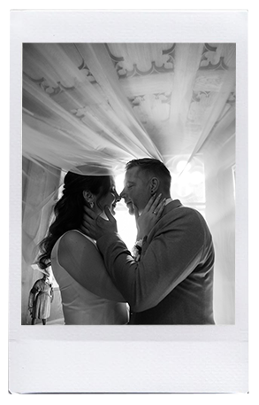 Braut und Bräutigam umarmen sich eng, lächeln und berühren sich, unter einer dekorativen Decke mit sanftem Licht, das durch ein Fenster strömt - wunderschön verziert mit moderner Hochzeitsfloristik von Dominique Kubicek Event & Floral Design Hochzeit.