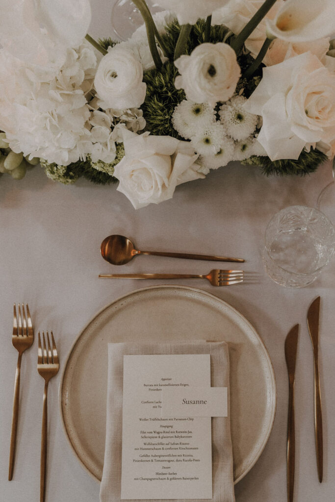 Ein stilvoll gedeckter Tisch mit einem beigen Teller, goldenem Besteck, einer Menükarte mit der Aufschrift Susanne, einem Glas und moderner Hochzeitsfloristik mit weißen Blumen und Grünzeug auf einer weißen Tischdecke.