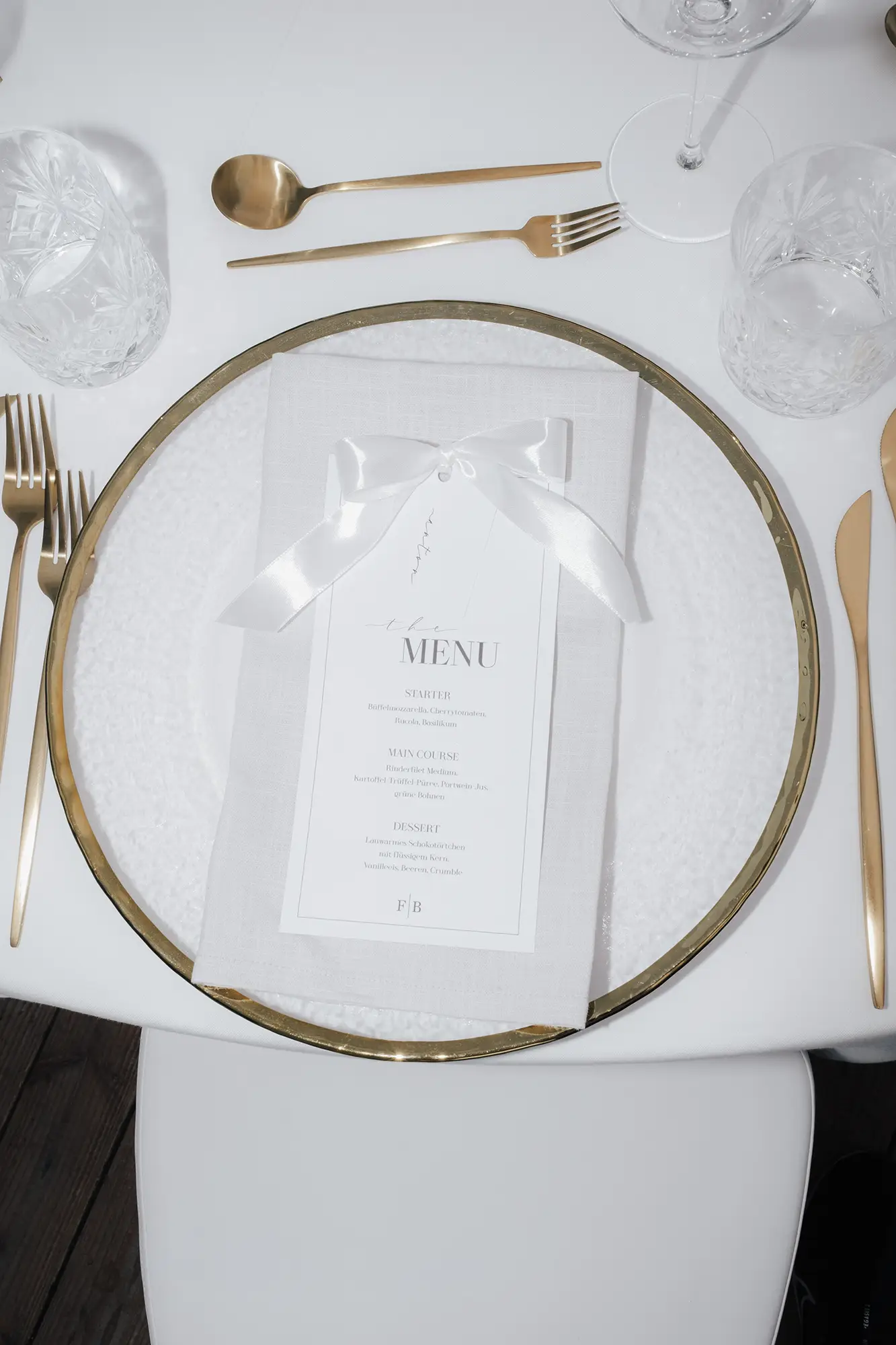 Ein formeller Tisch mit weißen Tellern auf einem goldverzierten Ladegerät, goldenem Besteck und Kristallgläsern spiegelt die moderne Hochzeitsfloristik von Event & Floral Design Hochzeit wider. Jede Menükarte wird elegant auf einer weißen Serviette zusammengebunden und sorgt für mondäne Raffinesse.