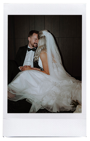 Eine Braut in weißem Kleid und Schleier sitzt eng mit einem Bräutigam im schwarzen Smoking zusammen. Umgeben von der modernen Hochzeitsfloristik von Dominique Kubicek lächeln sie, halten sich an den Händen und schauen sich liebevoll vor einem dunklen Hintergrund an.