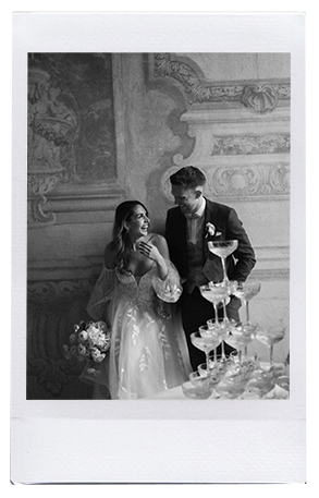Braut und Bräutigam lächeln neben einem gestuften Sektturm, umgeben von moderner Hochzeitsfloristik und kunstvollem Wandschmuck. Die Braut hält einen Blumenstrauß in der Hand und trägt ein fließendes Kleid, der Bräutigam trägt einen Anzug - und versprüht den Charme Niederbayerns.
