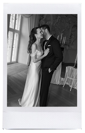 Eine Braut in einem weißen Kleid lächelt, während sie neben einem Bräutigam im Anzug steht. Aufgenommen von Dominique Kubicek, umarmen sie sich in der Nähe eines großen Fensters in einem eleganten niederbayerischen Raum mit Kerzen und kunstvoller Dekoration im Hintergrund.