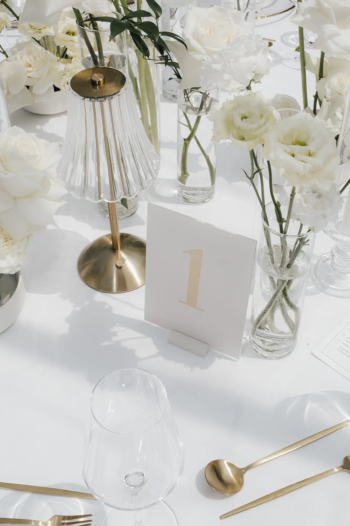 Elegante Tischdekoration mit weißen Blumen in klaren Vasen von Event & Floral Design Hochzeit, einer goldenen Lampe, goldenem Besteck, einem Weinglas und einer weißen Tischnummernkarte mit der Zahl 1 auf einer weißen Tischdecke - perfekt für moderne Hochzeitsfloristik in Niederbayern.