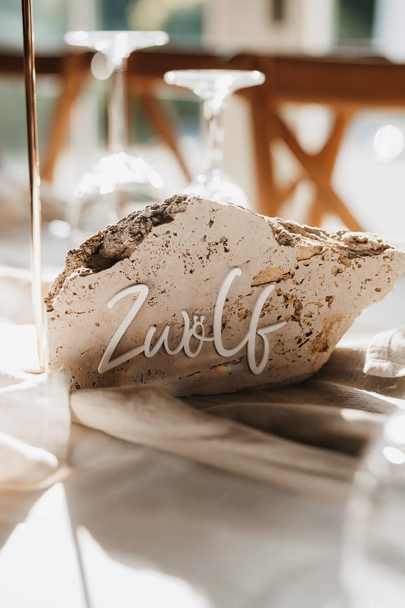 Ein rauer Stein mit dem Wort zwölf in weißer Schrift ruht auf einem mit leichtem Stoff drapierten Tisch, umgeben von verschwommenen Gläsern und in Sonnenlicht getaucht - eine heitere Szene, inspiriert von moderner Hochzeitsfloristik aus Niederbayern, gestylt von mondhaende.